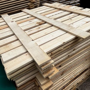 May include: A stack of unfinished wooden planks, likely for construction or crafts. The planks are light brown and arranged in neat rows, with a few planks laid across the top for stability. The wood grain is visible.
