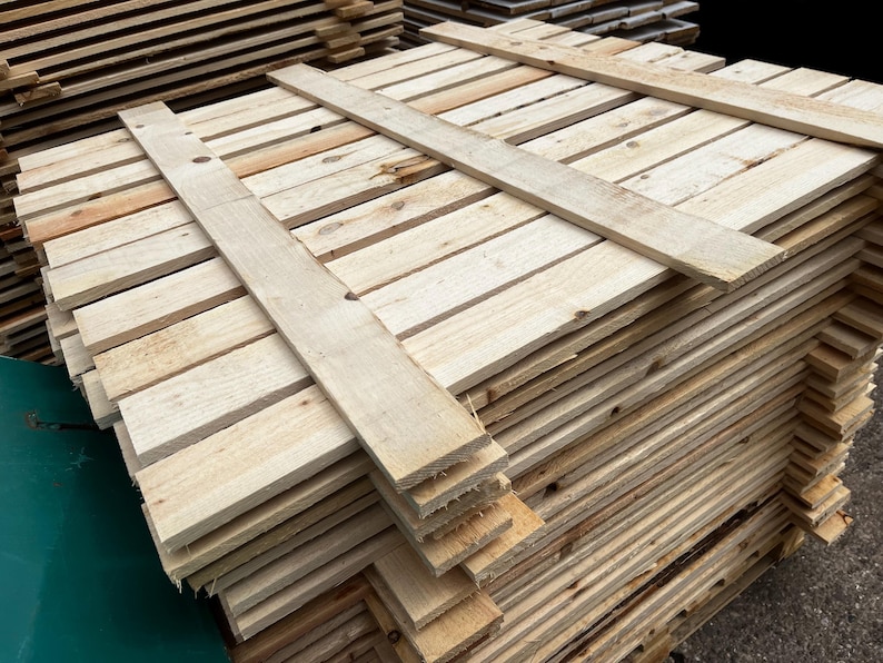 May include: A stack of unfinished wooden planks, likely for construction or crafts. The planks are light brown and arranged in neat rows, with a few planks laid across the top for stability. The wood grain is visible.
