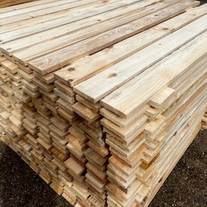 May include: A stack of unfinished wooden planks. The planks are light brown with visible wood grain and knots. The boards are of varying lengths and widths, creating a textured surface.