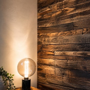 May include: A table lamp with a spherical glass bulb and a black marble base, emitting a warm glow. The lamp sits on a wooden surface next to a small potted plant, positioned against a wall featuring dark brown horizontal wooden planks.