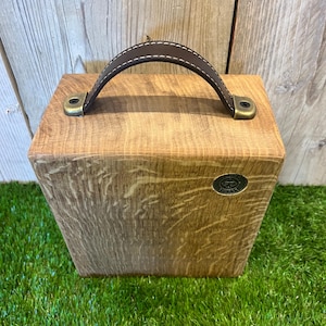 Puede incluir: Una caja rectangular de madera con acabado de veta natural, que cuenta con un asa de cuero marr&oacute;n oscuro sujeta con herrajes met&aacute;licos de color lat&oacute;n antiguo. Un peque&ntilde;o emblema met&aacute;lico circular con texto est&aacute; incrustado en la parte frontal.