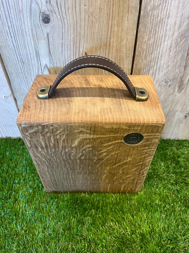 Puede incluir: Una caja rectangular de madera con acabado de veta natural, que cuenta con un asa de cuero marr&oacute;n oscuro sujeta con herrajes met&aacute;licos de color lat&oacute;n antiguo. Un peque&ntilde;o emblema met&aacute;lico circular con texto est&aacute; incrustado en la parte frontal.