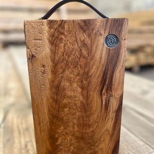 Puede incluir: Una tabla de servir rectangular de madera con un borde natural y un asa de cuero marr&oacute;n oscuro unida a la parte superior. La tabla presenta un patr&oacute;n de veta de madera prominente y una peque&ntilde;a insignia met&aacute;lica circular en la esquina superior derecha con el texto Funky Timber.