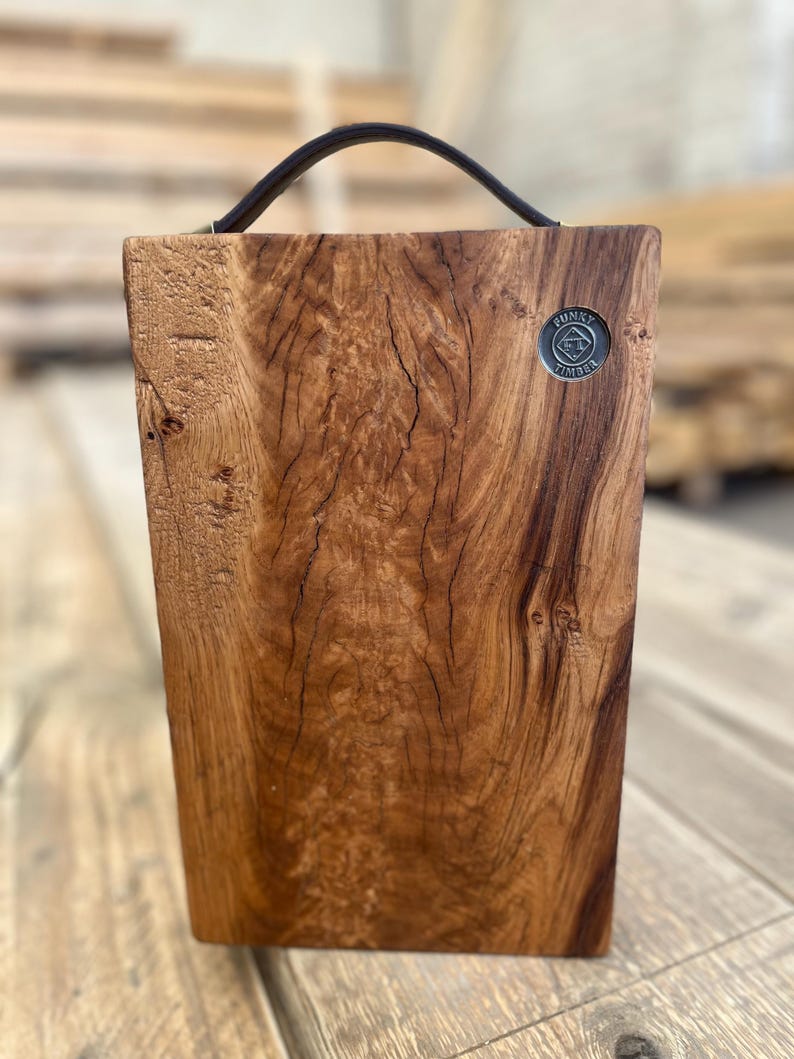 Puede incluir: Una tabla de servir rectangular de madera con un borde natural y un asa de cuero marr&oacute;n oscuro unida a la parte superior. La tabla presenta un patr&oacute;n de veta de madera prominente y una peque&ntilde;a insignia met&aacute;lica circular en la esquina superior derecha con el texto Funky Timber.