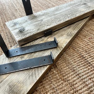 May include: Two rectangular wooden shelves with a light gray weathered finish and visible wood grain. Each shelf is paired with two L-shaped black metal brackets featuring pre-drilled holes for mounting, showcasing a raw industrial aesthetic.