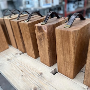 Puede incluir: Una serie de bloques rectangulares de madera dispuestos en fila, cada uno con un asa de cuero marr&oacute;n oscuro sujeta a la parte superior con herrajes met&aacute;licos de color lat&oacute;n. Los bloques son de madera natural con veta visible y acabado liso.