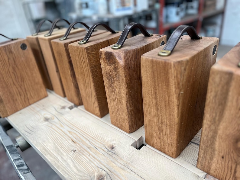 Puede incluir: Una serie de bloques rectangulares de madera dispuestos en fila, cada uno con un asa de cuero marr&oacute;n oscuro sujeta a la parte superior con herrajes met&aacute;licos de color lat&oacute;n. Los bloques son de madera natural con veta visible y acabado liso.