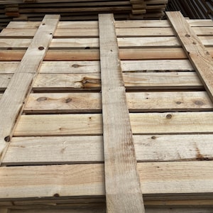 May include: Close-up of a wooden pallet made of light-colored planks. The pallet is constructed with horizontal boards and vertical supports, showing the natural wood grain and knots. The wood has a weathered appearance.