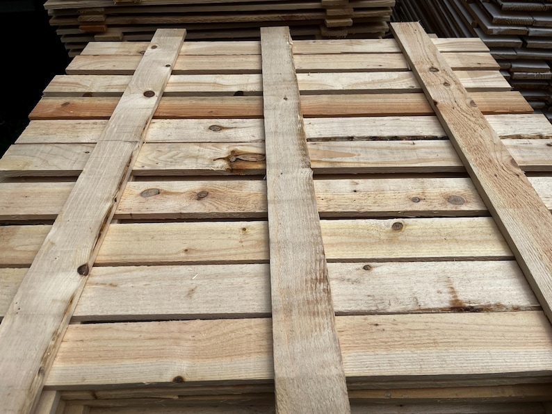 May include: Close-up of a wooden pallet made of light-colored planks. The pallet is constructed with horizontal boards and vertical supports, showing the natural wood grain and knots. The wood has a weathered appearance.