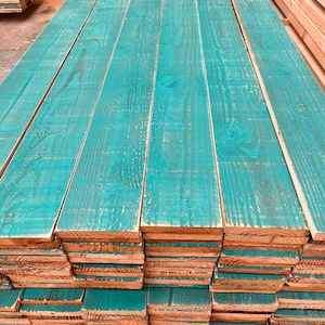 May include: A stack of teal-painted wooden planks. The planks are neatly stacked, showing the natural wood grain and texture. The teal paint has a distressed finish, adding character. The ends of the planks are a natural wood colour.