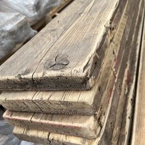 May include: A stack of four rectangular wooden planks with a weathered, greyish-brown finish. The boards feature visible wood grain, natural knots, and rough, textured edges with faint traces of red paint along the sides of the stacked lumber.