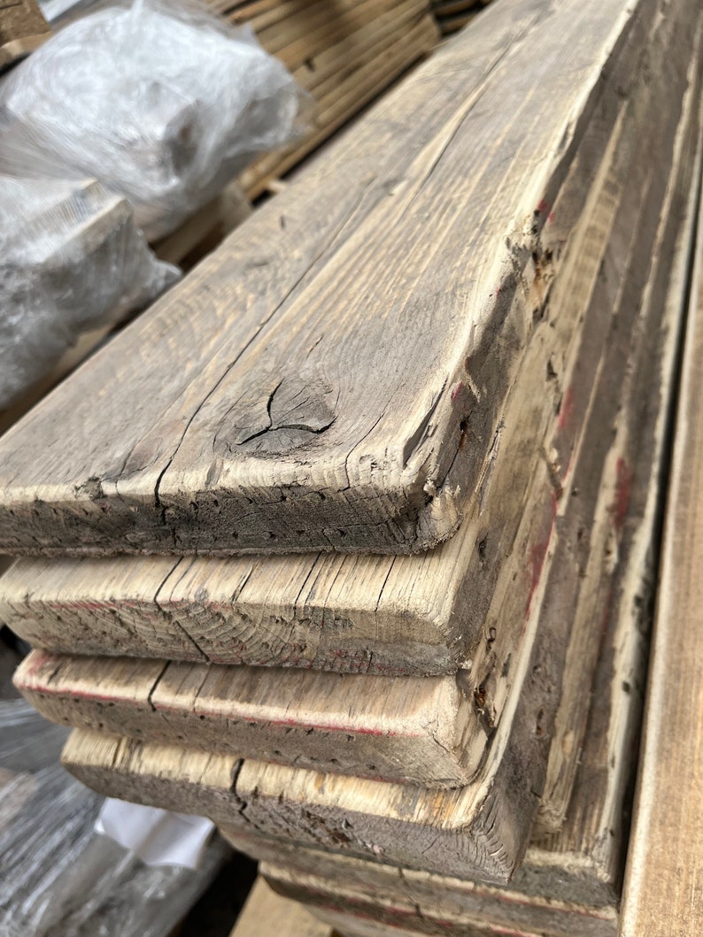 May include: A stack of four rectangular wooden planks with a weathered, greyish-brown finish. The boards feature visible wood grain, natural knots, and rough, textured edges with faint traces of red paint along the sides of the stacked lumber.