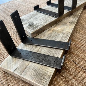 May include: Two rectangular light-colored wooden boards with visible grain patterns, each fitted with two L-shaped dark gray metal brackets. The metal brackets feature pre-drilled circular holes for mounting and have a textured, weathered finish.