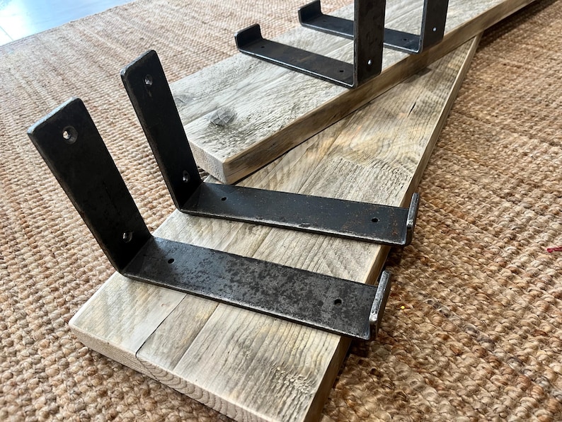 May include: Two rectangular light-colored wooden boards with visible grain patterns, each fitted with two L-shaped dark gray metal brackets. The metal brackets feature pre-drilled circular holes for mounting and have a textured, weathered finish.