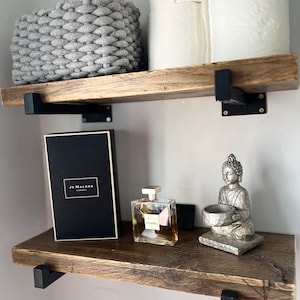 May include: Two rustic wooden shelves with black metal brackets. The top shelf holds a gray woven basket and two rolls of white toilet paper. The bottom shelf features a black Jo Malone box, a Chanel perfume bottle, and a silver Buddha figurine.