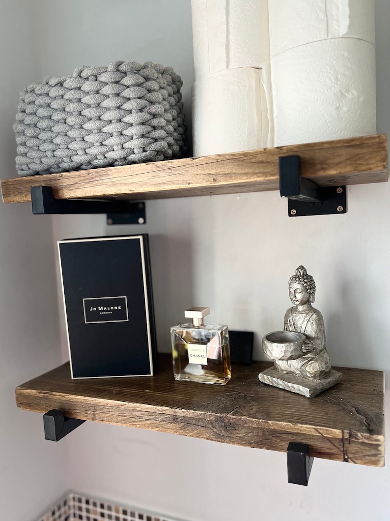 May include: Two rustic wooden shelves with black metal brackets. The top shelf holds a gray woven basket and two rolls of white toilet paper. The bottom shelf features a black Jo Malone box, a Chanel perfume bottle, and a silver Buddha figurine.
