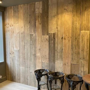 May include: A wooden wall constructed from vertical planks in shades of brown and grey. The wall serves as a backdrop for three ornate, black metal side tables. A window is visible on the left side of the image.