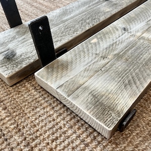 May include: Two rectangular wooden shelves featuring a weathered gray finish with visible wood grain patterns. Each shelf is attached to black metal brackets with pre-drilled holes for mounting, showcasing a flat surface and straight edges.