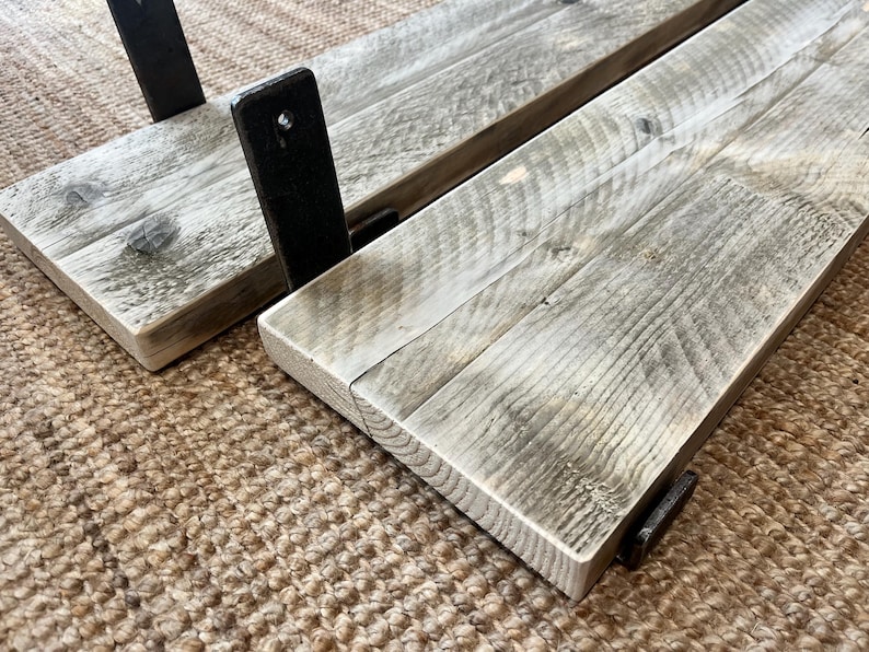 May include: Two rectangular wooden shelves featuring a weathered gray finish with visible wood grain patterns. Each shelf is attached to black metal brackets with pre-drilled holes for mounting, showcasing a flat surface and straight edges.
