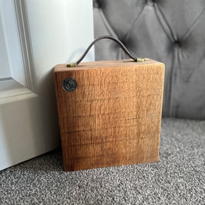 Puede incluir: Un bloque de madera cuadrado con veta visible, que cuenta con un asa de cuero marr&oacute;n oscuro sujeta con herrajes de color lat&oacute;n. Un peque&ntilde;o emblema met&aacute;lico circular con el texto "TONY'S TIMBER" est&aacute; incrustado en la esquina superior izquierda.