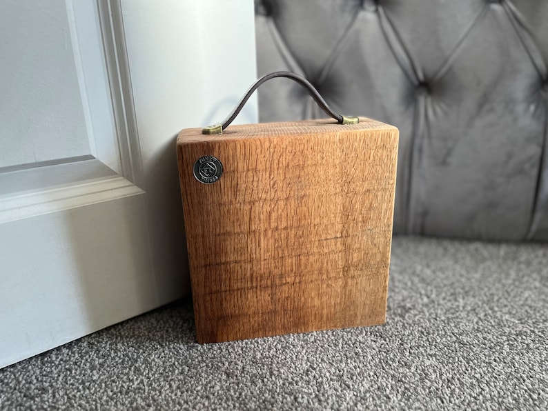 Puede incluir: Un bloque de madera cuadrado con veta visible, que cuenta con un asa de cuero marr&oacute;n oscuro sujeta con herrajes de color lat&oacute;n. Un peque&ntilde;o emblema met&aacute;lico circular con el texto "TONY'S TIMBER" est&aacute; incrustado en la esquina superior izquierda.