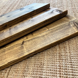 May include: Three rectangular wooden boards with a dark brown stained finish, showcasing visible wood grain patterns, knots, and a smooth, polished surface texture. The boards are arranged in a staggered, overlapping formation, highlighting the natural variations in the wood tone and the straight, clean edges of each individual plank.