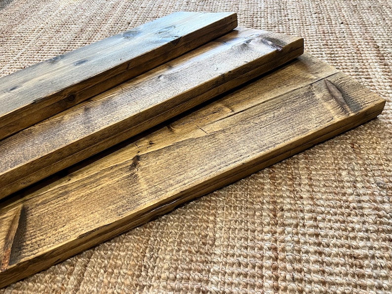 May include: Three rectangular wooden boards with a dark brown stained finish, showcasing visible wood grain patterns, knots, and a smooth, polished surface texture. The boards are arranged in a staggered, overlapping formation, highlighting the natural variations in the wood tone and the straight, clean edges of each individual plank.