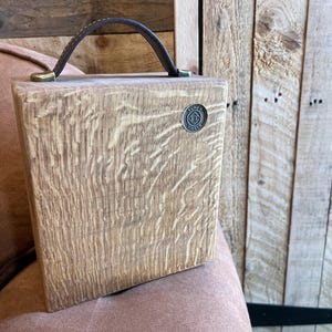 Puede incluir: Una caja cuadrada de madera con veteado marr&oacute;n claro, que cuenta con un asa de cuero marr&oacute;n oscuro sujeta con herrajes de color lat&oacute;n. Un peque&ntilde;o emblema met&aacute;lico circular est&aacute; incrustado en la esquina superior derecha con el texto Funky Timber.