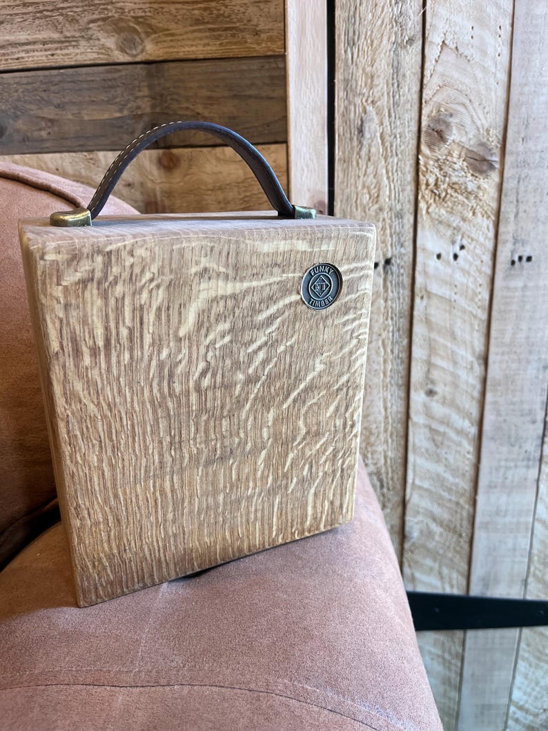 Puede incluir: Una caja cuadrada de madera con veteado marr&oacute;n claro, que cuenta con un asa de cuero marr&oacute;n oscuro sujeta con herrajes de color lat&oacute;n. Un peque&ntilde;o emblema met&aacute;lico circular est&aacute; incrustado en la esquina superior derecha con el texto Funky Timber.