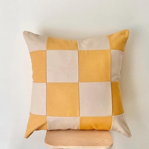 May include: Square decorative pillow with a checkerboard pattern in shades of yellow and cream. The pillow is made of a soft fabric and is resting on a wooden stool. The pillow's design adds a touch of modern style to any room.