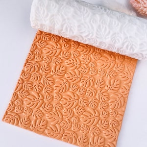 Puede incluir: Un rodillo de silicona blanco con un patrón de hojas y una hoja de silicona naranja a juego con el mismo patrón de hojas.