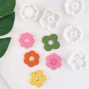 Puede incluir: Surtido de nueve cortadores de galletas con forma de flor en varios colores, incluyendo blanco, rosa, amarillo, naranja y verde. Los cortadores están dispuestos sobre una superficie blanca.