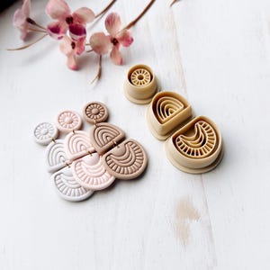 May include: A selection of beige polymer clay earring components and finished earrings in various shapes, including circles, arches, and half-circles. The earrings are in shades of white, pink, and beige. A sprig of pink flowers is in the background.