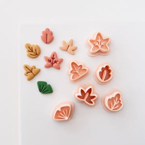Könnte beinhalten: Eine Sammlung von hellpfirsichfarbenen Kunststoff-Ausstechformen in Blattform sowie mehrere kleine, fertige Blattformen in gedeckten Orange-, Gelb-, Rosa- und Grüntönen, die auf einer weißen Oberfläche zum Backen oder Basteln angeordnet sind.