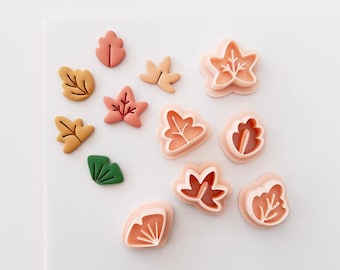 Può includere: Una collezione di tagliabiscotti in plastica color pesca chiaro a forma di foglia insieme a diverse piccole forme di foglie finite in tonalità tenui di arancione, giallo, rosa e verde, disposte su una superficie bianca per dolci o artigianato.