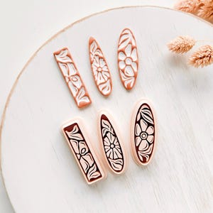 Op de afbeelding: Een set van zes klei-uitsteekvormpjes in rechthoekige, druppelvormige en ovale vormen. De bovenste rij bevat terracotta-kleurige uitsteekvormpjes met witte bloem- en bladreliëfpatronen. De onderste rij bevat lichtroze uitsteekvormpjes met donkerbruine bloemmotieven.