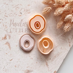 Peut inclure: Trois emporte-pièces en argile de forme circulaire irrégulière dans des tons pêche et crème. Les outils présentent une découpe ovale centrale et un bord extérieur surélevé. Le texte "Paulas CUTTERS" est visible sur le côté gauche.