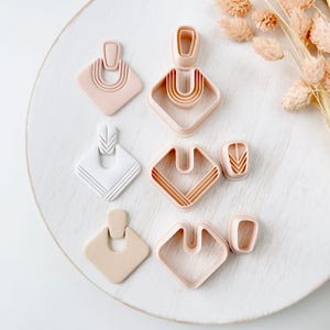 Peut inclure: Un ensemble de trois boucles d'oreilles géométriques finies en rose poudré, blanc et beige, accompagné de trois emporte-pièces en plastique rose clair assortis. Les boucles présentent des textures en arche et chevron superposées.