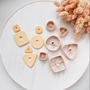 May include: A collection of light beige textured clay shapes including squares, circles, and triangles, alongside several matching pale pink plastic cutters in various geometric forms, arranged on a white circular surface.