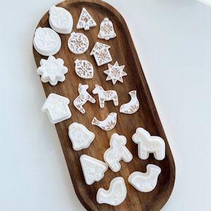 Puede incluir: Una colección de adornos y formas navideñas en blanco y dorado, que incluyen estrellas, campanas y casas, dispuestas en una bandeja de madera. Los adornos presentan patrones y diseños intrincados, perfectos para la decoración navideña.