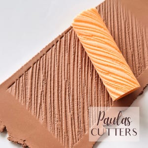 May include: Close-up of a textured clay surface with a diagonal pattern created by a rolling pin. The rolling pin is orange and has a wavy pattern. The clay is a light brown color. The text "Paulas CUTTERS" is in the bottom right corner.