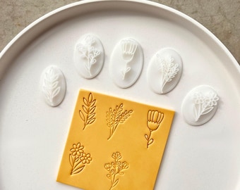 Puede incluir: Un conjunto de cinco sellos blancos de forma ovalada con diseños botánicos en relieve y una lámina de arcilla cuadrada amarilla con cinco impresiones florales correspondientes, incluyendo varios patrones de hojas, tallos y flores.