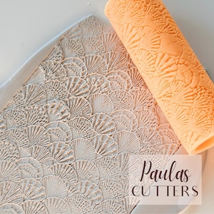Può includere: Una vista dall'alto mostra un mattarello strutturato e un foglio di argilla con un motivo a ventaglio. Il mattarello è arancione e l'argilla è di colore beige chiaro. Il testo "Paulas CUTTERS" è in un rettangolo bianco.
