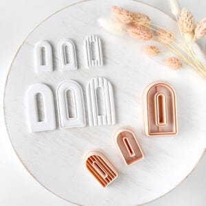 May include: A collection of arch-shaped clay cutters in various sizes, featuring both solid white and light peach plastic designs. Some cutters have smooth surfaces, while others display vertical ribbed textures for creating detailed shapes.