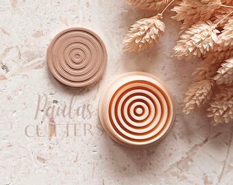May include: A light peach-colored circular clay cutter featuring a concentric ring pattern on its face, accompanied by a matching circular stamp with a similar embossed ring design, with the text Paulas CUTTERS visible in the background.