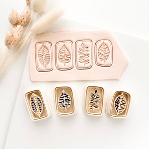 May include: Four leaf-shaped clay stamps with different vein patterns are displayed. The stamps are in a rectangular shape with a white border. A piece of pink clay with the same leaf designs is also shown. The background is white.