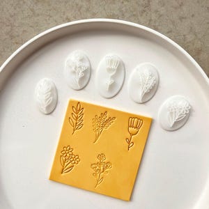 Peut inclure: Ensemble d'outils artisanaux sur le thème floral. L'image présente six tampons blancs de forme ovale avec divers motifs floraux, ainsi qu'un carré jaune avec des motifs floraux en relief. Les articles sont disposés sur un plateau blanc et rond.