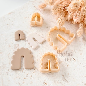 May include: A collection of beige-colored cookie cutters in the shape of rainbows. The cutters have a scalloped edge and are made of plastic. The image is taken from a top-down perspective, with the cutters arranged on a light-colored surface.