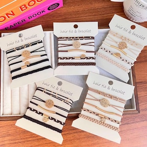 May include: Five sets of hair tie and bracelet accessories displayed on white cards. Each set includes multiple bands in black, brown, beige, or tan, featuring gold-toned metal accents, circular filigree charms, and braided textures.