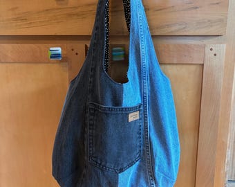 May include: A hobo-style shoulder bag crafted from two-toned denim, featuring a dark charcoal grey panel on the left and a lighter blue denim panel on the right. The bag includes a front patch pocket with a small rectangular label and a black and white polka dot interior lining visible at the top opening.
